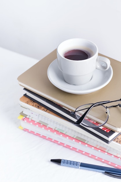 Stack of different books and notepads with woman's glasses and coffee cup on the top on a white table