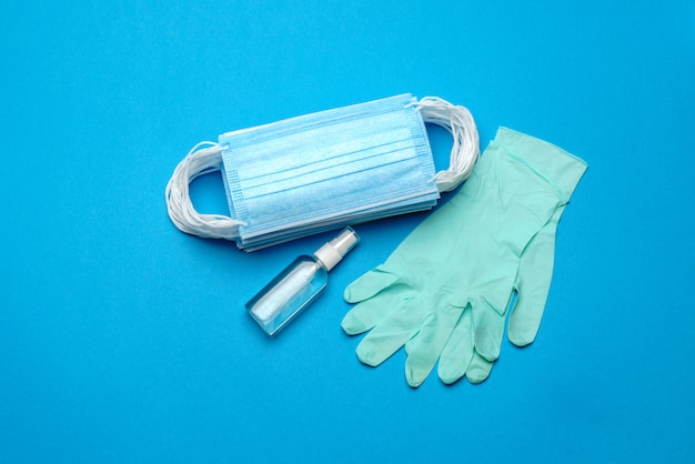 Stack of Disposable blue medical face masks, rubber latex gloves and alcohol hand sanitizer antiseptic on blue wall