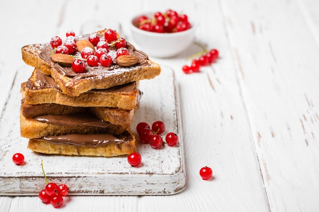 Stack of fried toast with chocolate cream, red currant and almonds on a white wooden background