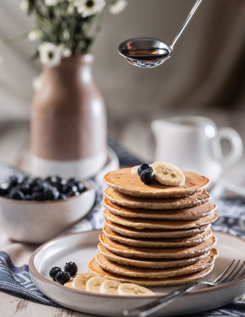 Stack of home baked banana pancakes with blueberries, gravy boat with maple syrup hangs over pancakes