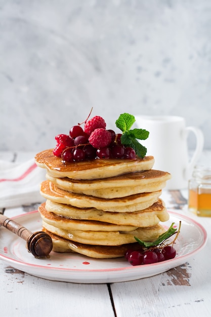 Stack of homemade little pancakes with honey, fresh raspberries and red currants on an old  light wooden surface.