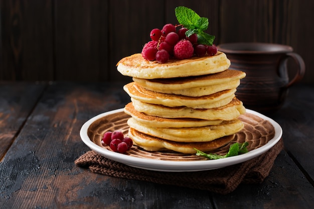 Stack of homemade little pancakes with honey, fresh raspberries and red currants on an old wooden surface.