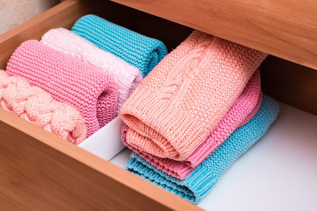 A stack of knitted clothes next to a box of neatly folded items in a dresser drawer.