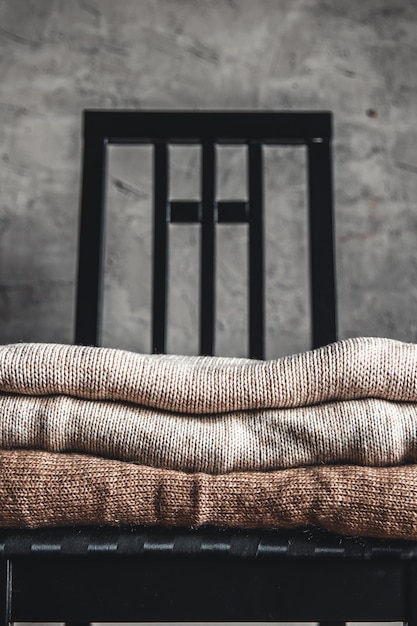 Photo a stack of knitted warm cozy sweaters , on a chair by the gray wall. autumn, winter concept.