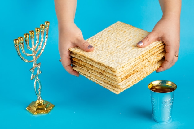 A stack of matzo in female hands on a blue surface next to a menorah glass of wine