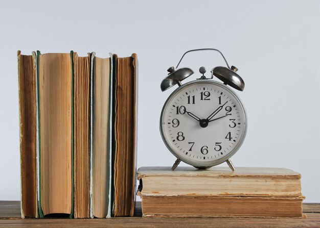 Stack of old books and alarm clock on woden shelf against the white wall