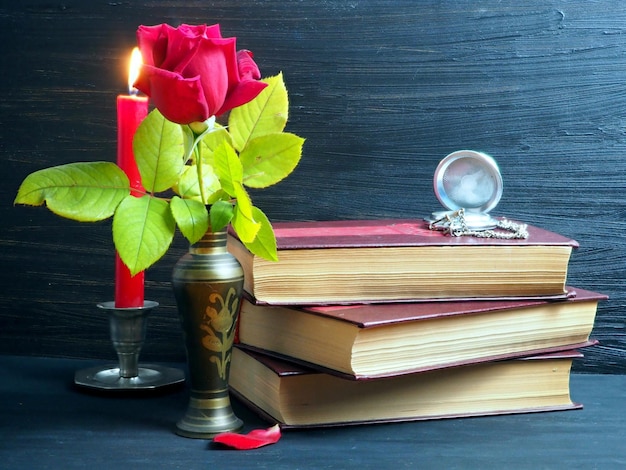 A stack of old books an old pocket watch a burning candle a red rose in a vase