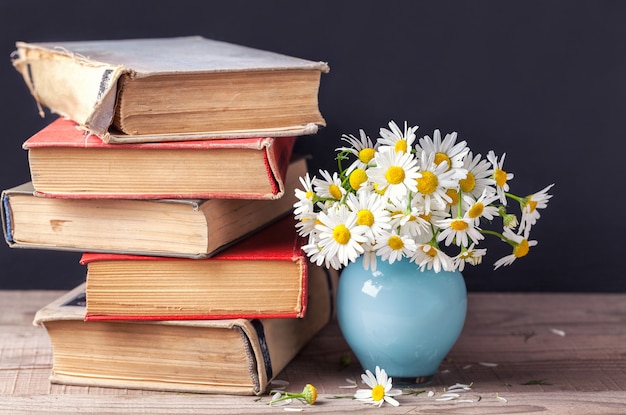 A stack of old vintage books lying on a wooden shelf with a bouquet of daisies in a blue vase. 