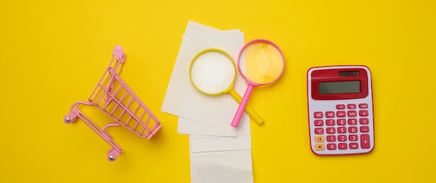 Stack of paper checks, a pink plastic calculator and a magnifying glass on a yellow background. Family budget audit concept, search for savings