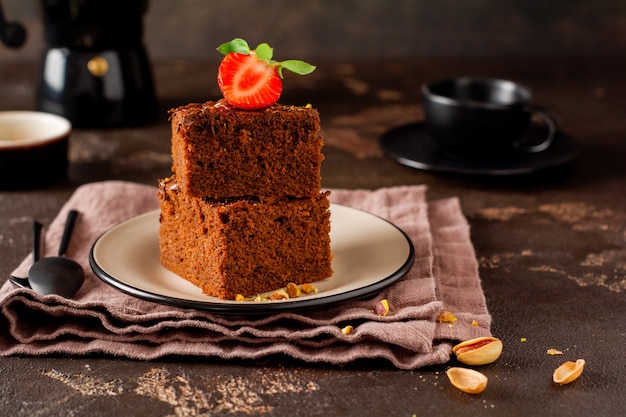 Stack of pieces or bar of chocolate cake brownie with strawberries and pistachio nuts on the black surface, selective focus image.