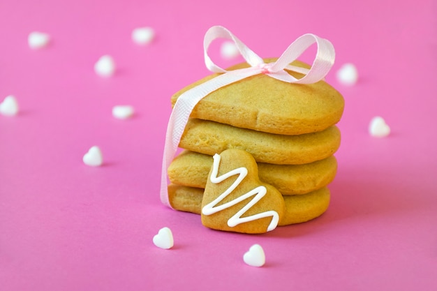 Stack of shortbread cookies tied with pink ribbon and many white heart shape candy confetti on pastel pink surface. Freshly baked homemade bakery, tasty sugar biscuits, teatime.