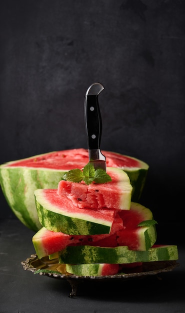 Stack of slices of ripe red watermelon in a metal plate, stuck in a large knife. Black background