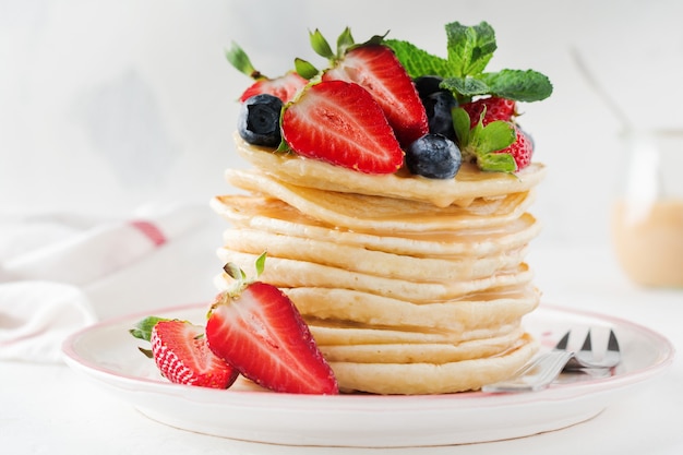 Stack of small pancakes for breakfast with strawberries, blueberries and sweet sauce on a light table. Selective focus.