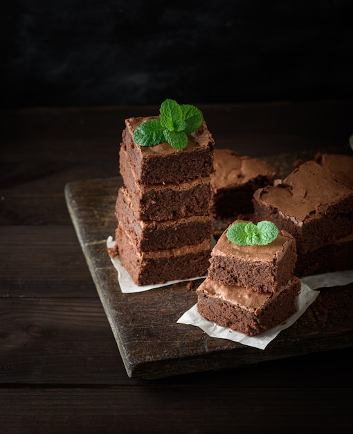 Stack of square pieces of baked brown brownie pie on a wooden  board, on the top is a green mint leaf