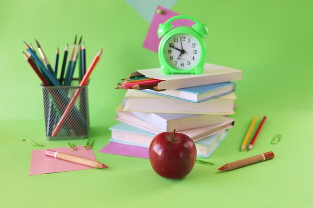 Stack of textbooks, alarm clock, apple and school supplies, green background