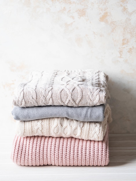 Stack of warm knitted woolen sweaters pastel shades against beige textured wall. Copy space