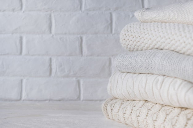 Stack of white cozy knitted sweaters against the background of white brick wall.