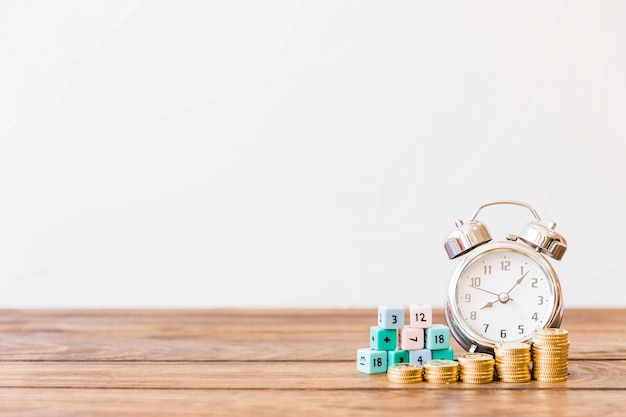 Stacked coins, alarm clock and math blocks on wooden surface