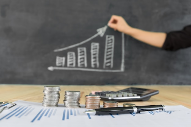 Stacked of coins on desk with person hand showing growth chart on blackboard