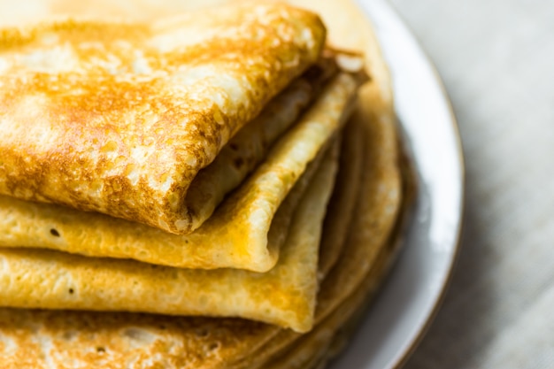 Photo stacked folded crepes on white plate on linen cloth closeup, breakfast