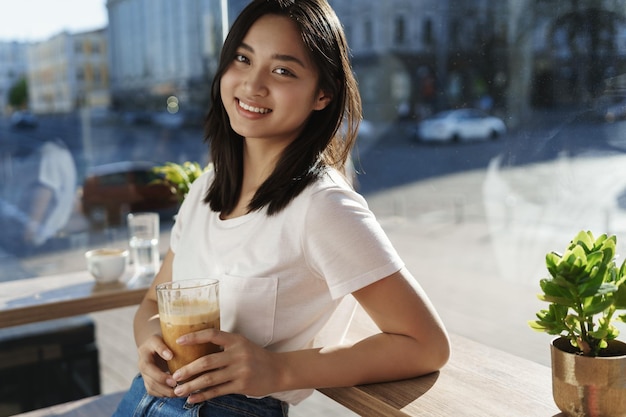 Stad en jongeren nieuwe generatie concept Aziatische lachende vrouw in favoriete café magere tafel in de buurt van raam achter straten vrouw houdt koffiecocktail en kijk camera vreugdevol ontmoeting vriend