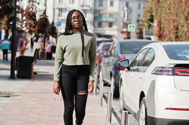 Stadsportret van positief jong donker gevild wijfje groen hoody dragen en oogglazen die bij autoparkeren lopen.