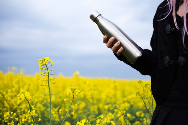 Stainless thermos bottle in hand, on surface of rapeseed field.