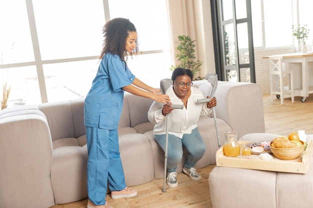 Stand from sofa. Caring nurse helping woman with arthritis to stand from the sofa with crutches