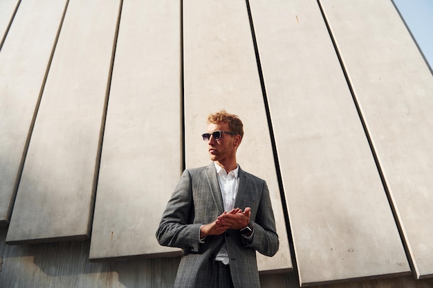 Standing against building wall Young businessman in grey formal wear is outdoors in the city