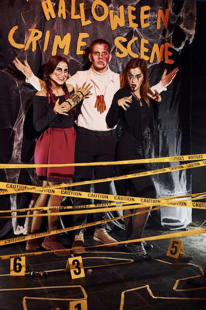 Standing against crime scene wall. Friends is on the thematic halloween party in scary makeup and costumes.