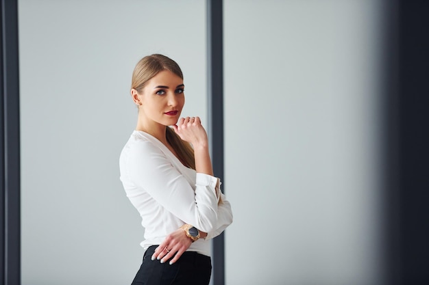 Standing against grey background Young adult woman in formal clothes is indoors in the office