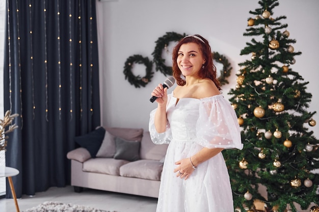 Standing in the christmas decorated domestic room woman in\
white dress and with microphone in hands is singing in the