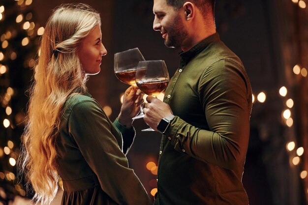 Standing and holding big glasses with wine Young lovely couple have romantic dinner indoors together