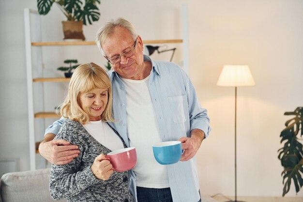 Standing and holding cups of drinks senior man and woman is\
together at home