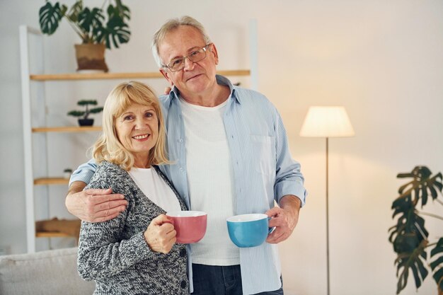 Standing and holding cups of drinks senior man and woman is\
together at home