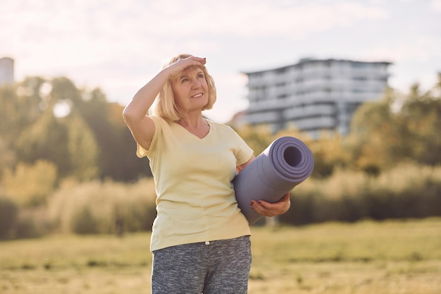 Standing and holding yoga mat senior woman having nice weekend\
outdoors on the field at sunny day