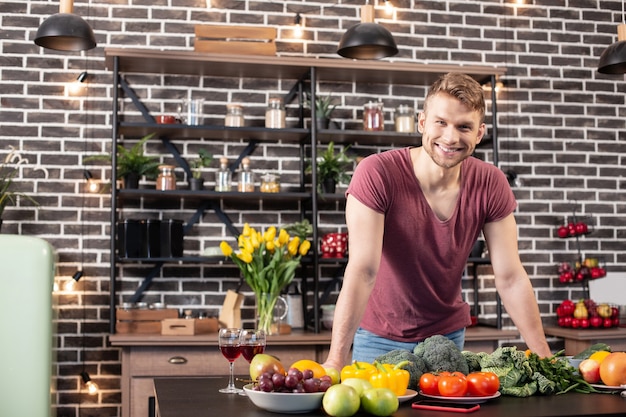 In piedi in cucina. bel ragazzo barbuto che sorride ampiamente mentre si trova in cucina prima di cucinare