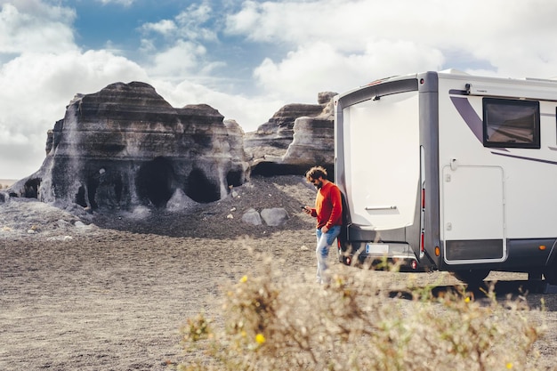 Standing man using mobile phone against a modern camper van and\
country side rural landscape in background concept of travel and\
connection technlogy to plan road trip and holiday vacation