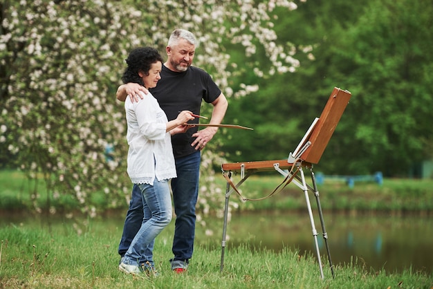 Standing near the lake. Mature couple have leisure days and working on the paint together in the park