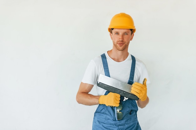 Standing near white wall Young man working in uniform at construction at daytime