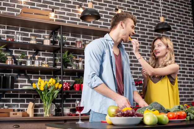 Standing near wife. Bearded handsome caring husband cutting vegetables for salad standing near wife