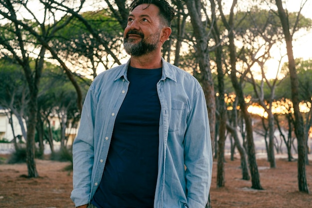 Standing portrait of man in casual clothes enjoying time in outdoor leisure activity at the park Trees in background Tourist concept Mature male people smile and look in the air Nature around