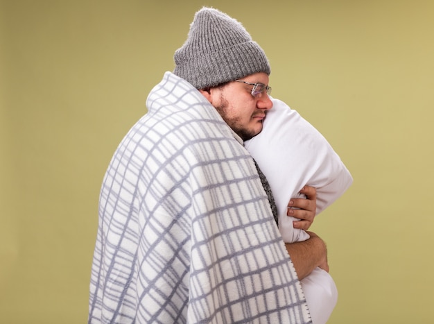 Standing in profile view with closed eyes middle-aged ill male wearing winter hat and scarf wrapped in plaid hugged pillow 