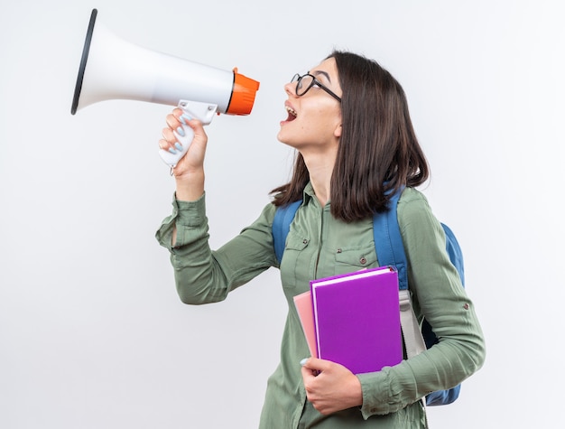 Standing in profile view young school woman wearing glasses with backpack holding books speaks on loudspeaker 