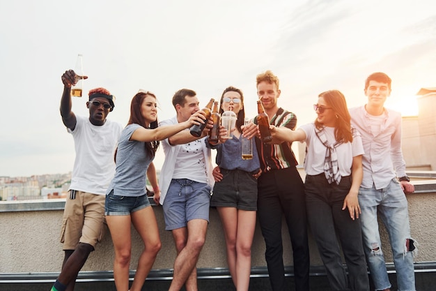 Standing with alcohol at the edge of rooftop Group of young people in casual clothes have a party together at daytime