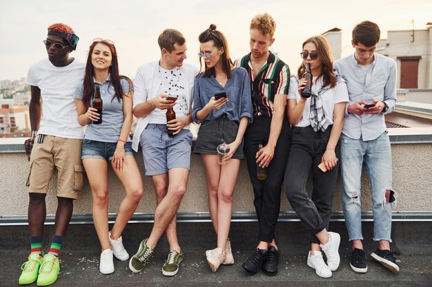 Photo standing with alcohol at the edge of rooftop group of young people in casual clothes have a party together at daytime