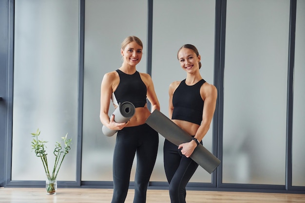 Стоя с ковриками в руках Две женщины в спортивной одежде и со стройными телами вместе проводят день фитнес-йоги в помещении