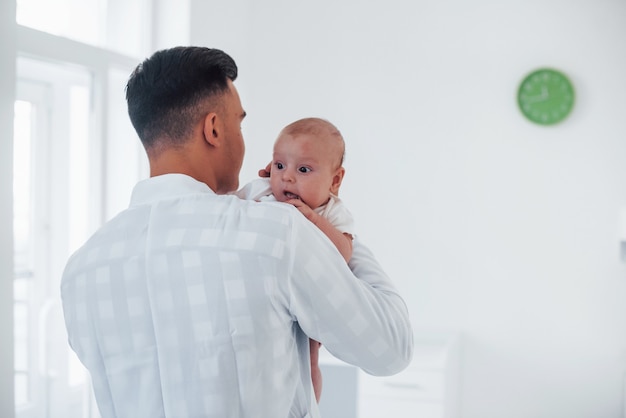 Stands and holds baby on the hands. Young pediatrician is in the clinic at daytime.