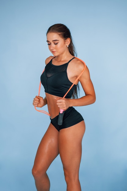 Stands with jumping rope. Young woman with slim body type isolated against blue background.