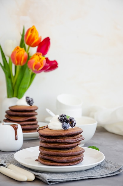 Stapel chocolade pannenkoeken op een witte plaat met chocoladesaus, slagroom, bosbessen en bramen bovenop een betonnen achtergrond.